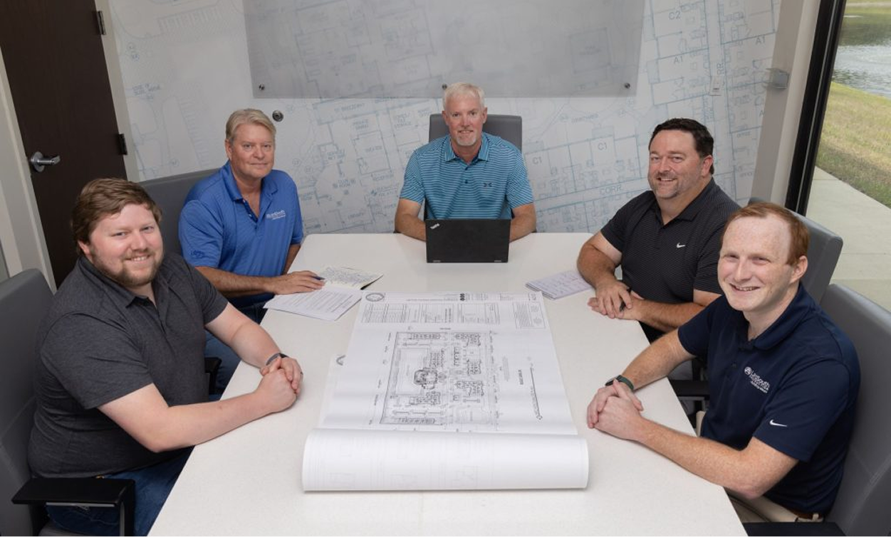 Five men sit around a conference table with building plans and documents, meeting in an office with architectural drawings on the wall—an ideal setting for multifamily general contractors discussing market rate developments.
