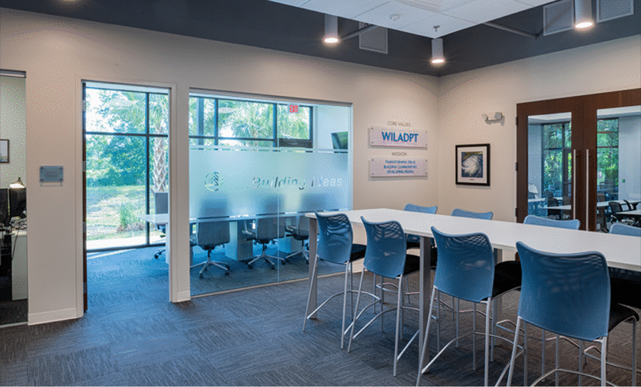 Modern office space with blue chairs around a white table, glass-walled conference room, and large windows letting in natural light—ideal for land development or market rate developments teams to collaborate and innovate.
