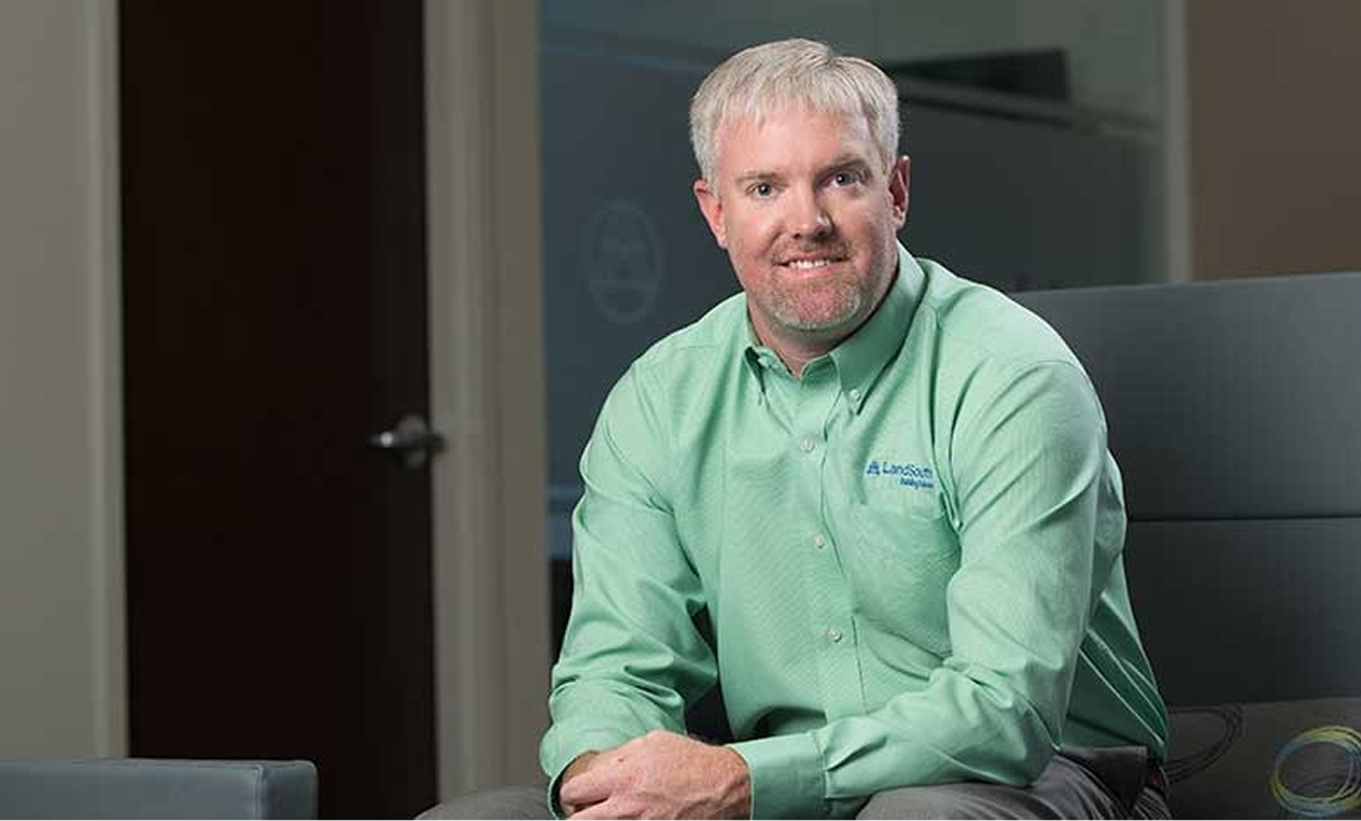 A man with short light hair and a beard, wearing a green button-up shirt, sits indoors on a chair, facing the camera with a neutral expression—reflecting the thoughtful approach often required in land development projects.