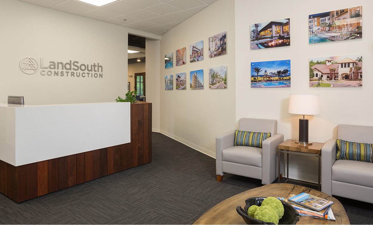 Modern office reception area with a white and wood desk, two armchairs, a side table with a lamp, wall art featuring building photos, and the LandSouth Construction logo—highlighting expertise as multifamily general contractors.